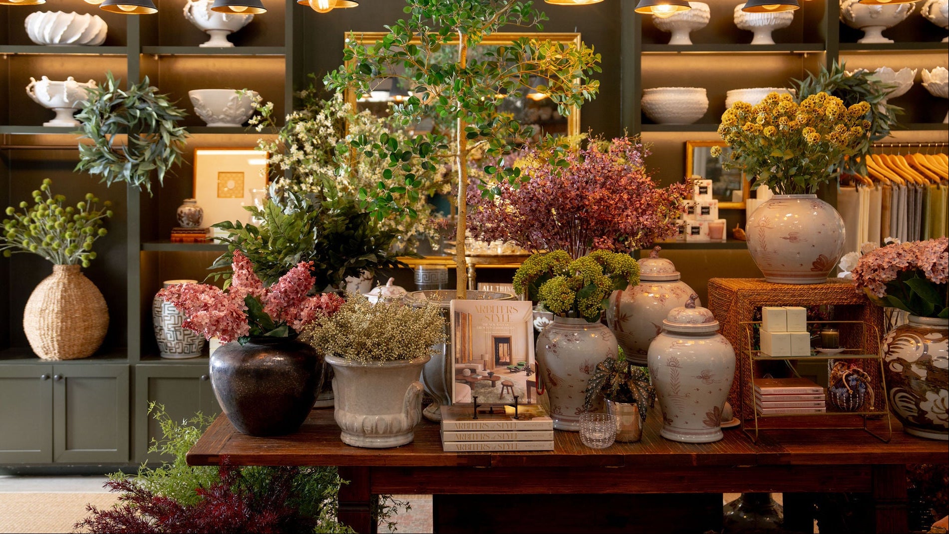 A beautifully styled interior room featuring a large olive green shelving unit filled with ceramic vases, bowls, and potted greenery, alongside a wooden table displaying multiple floral arrangements in textured ceramic pots under hanging black pendant lights.